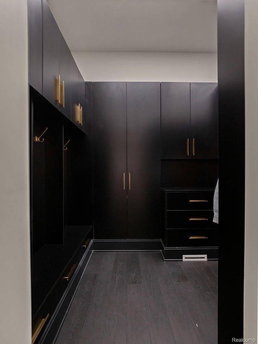 This is a modern walk-in closet featuring dark cabinetry with gold hardware and dark hardwood flooring. The closet includes ample storage with both hanging space and drawers. The overall impression is luxurious and well-organized.