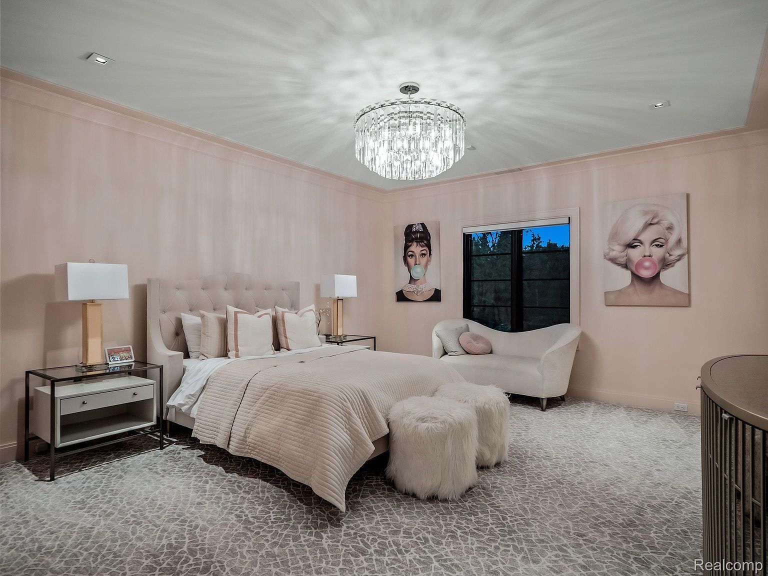 This is a luxurious primary bedroom featuring a tufted headboard bed with neutral bedding, flanked by glass-topped nightstands and lamps. A crystal chandelier illuminates the room, and artwork depicting iconic figures adds a touch of glamour. The room also includes a chaise lounge and plush ottomans, creating a comfortable and stylish retreat.
