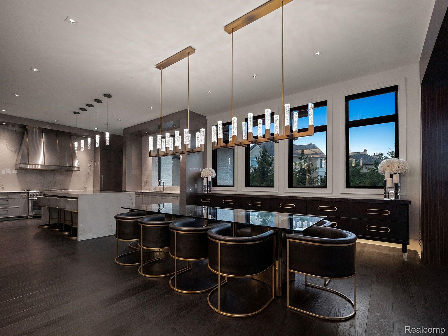 This is an interior shot of a modern dining room featuring a long, black dining table with black and gold chairs. Above the table are three modern chandeliers, and large windows provide natural light and a view of the outdoors. The dark wood flooring and sleek cabinetry add to the room's contemporary and luxurious feel.