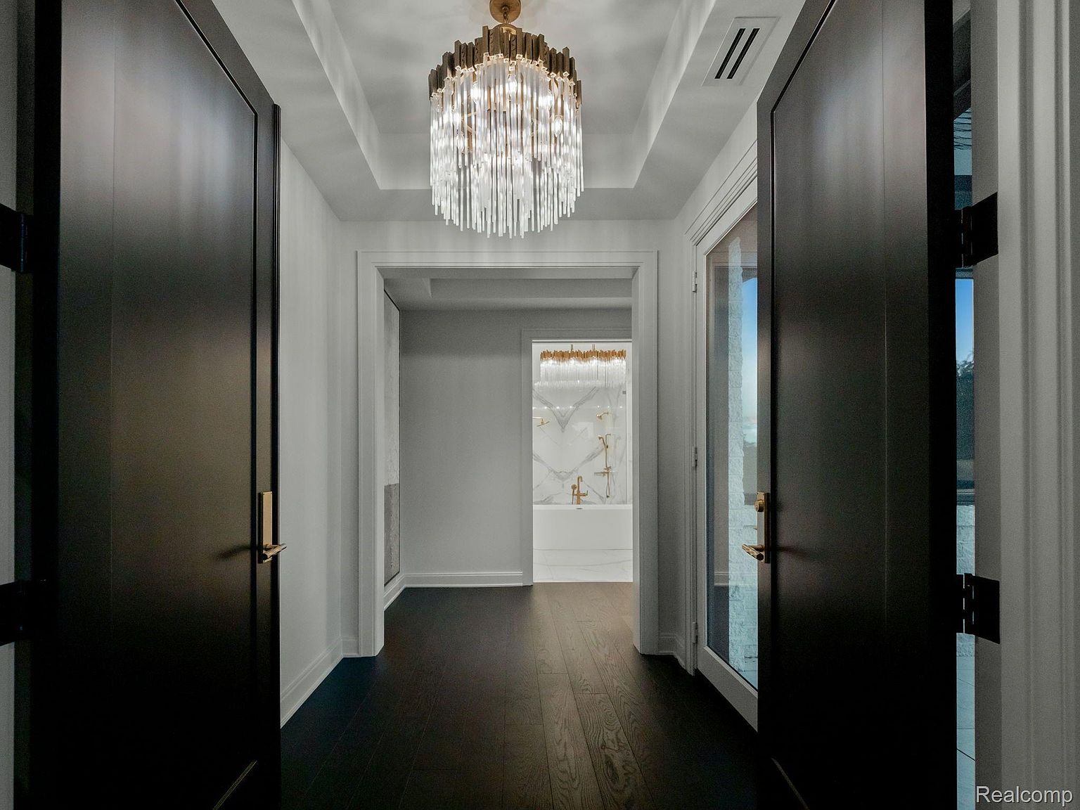 This interior shot showcases a luxurious hallway leading to a bathroom. The hallway features dark hardwood floors, white walls, and a striking crystal chandelier. Two dark doors frame the view, adding depth and elegance to the space, while the bathroom in the background features a freestanding tub and gold fixtures.