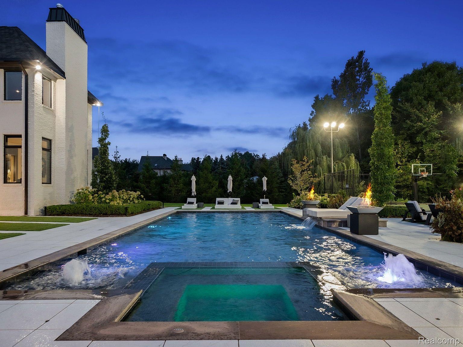 This image showcases a luxurious outdoor pool and spa area at dusk. The pool features integrated lighting, fountains, and a separate spa section, all surrounded by a spacious patio. Fire features and lounge seating enhance the ambiance, creating an inviting and upscale outdoor living space.