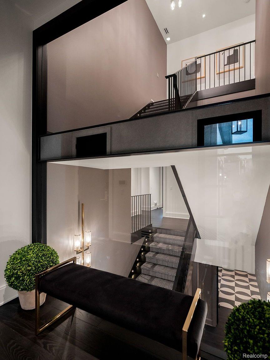 This interior shot showcases a modern hallway and staircase with a unique architectural design. The space features a combination of dark wood flooring, carpeted stairs with a geometric pattern, and glass partitions, creating a visually striking effect. A black velvet bench with gold accents adds a touch of luxury to the entryway.