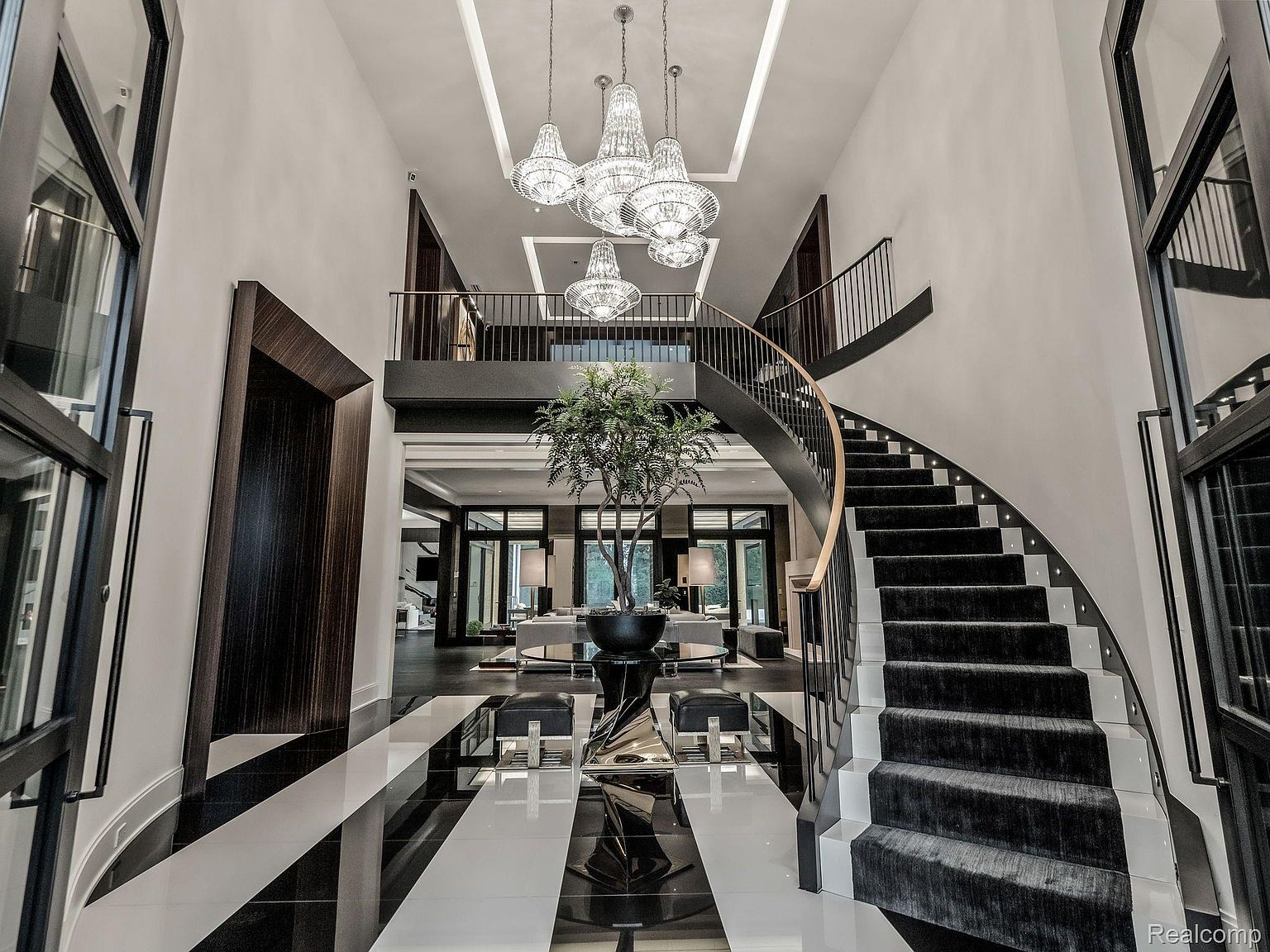 This grand foyer features a striking black and white striped tile floor, a curved staircase with dark carpeting, and a large potted tree as a focal point. Multiple crystal chandeliers hang from the high ceiling, adding to the luxurious ambiance. The view is from the entrance, looking into the living space beyond.