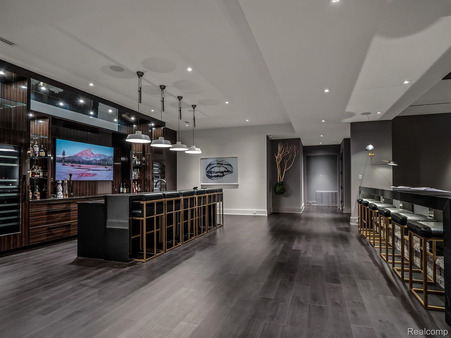 This is a modern basement featuring a bar area with dark wood floors and a black and gold bar. The bar is equipped with seating and pendant lighting, and there is a large television mounted on the wall. The space is well-lit with recessed lighting and has a contemporary design.