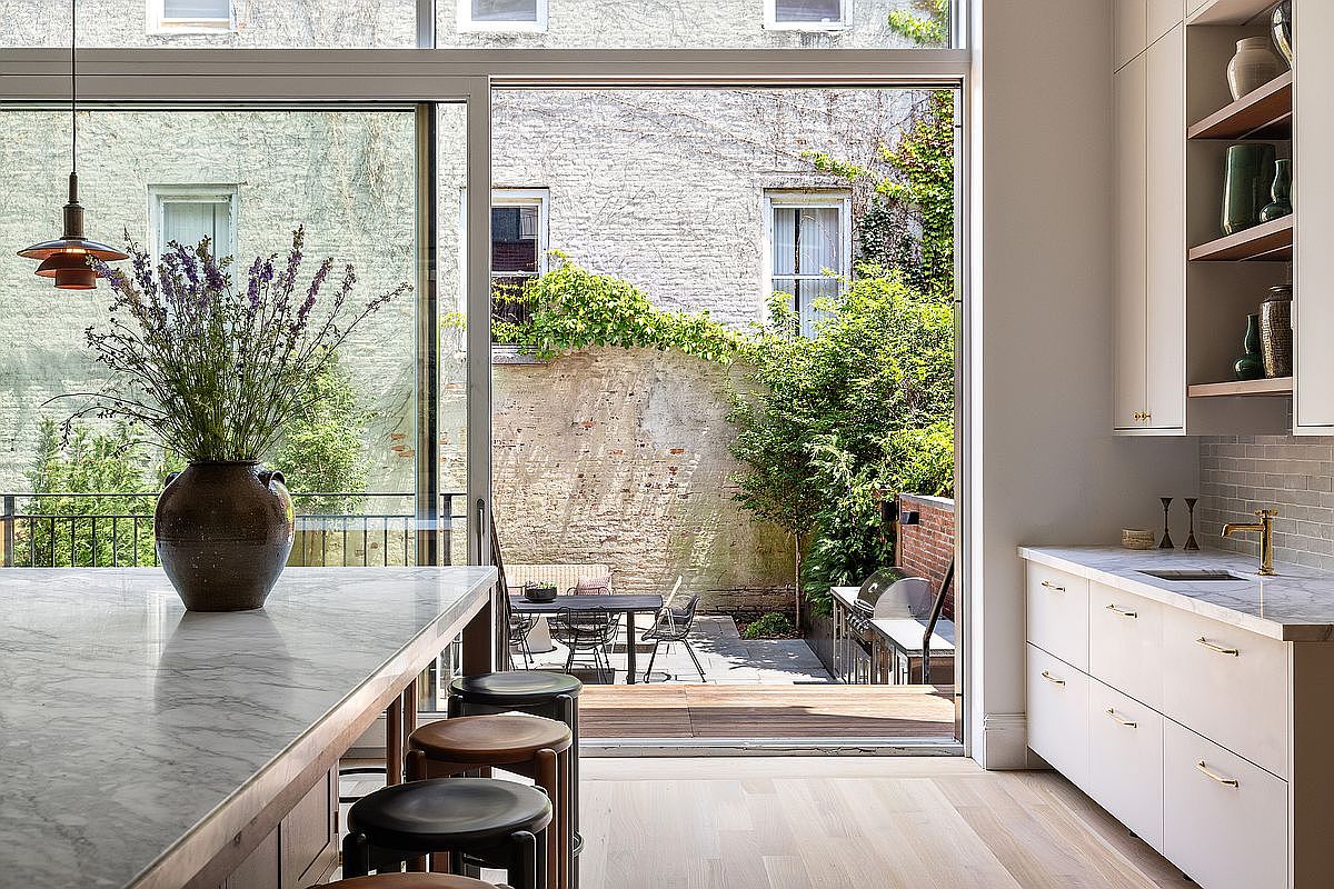 This bright and airy kitchen features a large marble-topped island with dark wooden stools, seamlessly connecting to an outdoor patio through expansive floor-to-ceiling sliding glass doors. The space is characterized by clean, minimalist white cabinetry, open shelving, and a warm, natural wood floor that creates a sophisticated, modern aesthetic. The view through the glass highlights a lush, private courtyard, emphasizing a harmonious indoor-outdoor living experience.