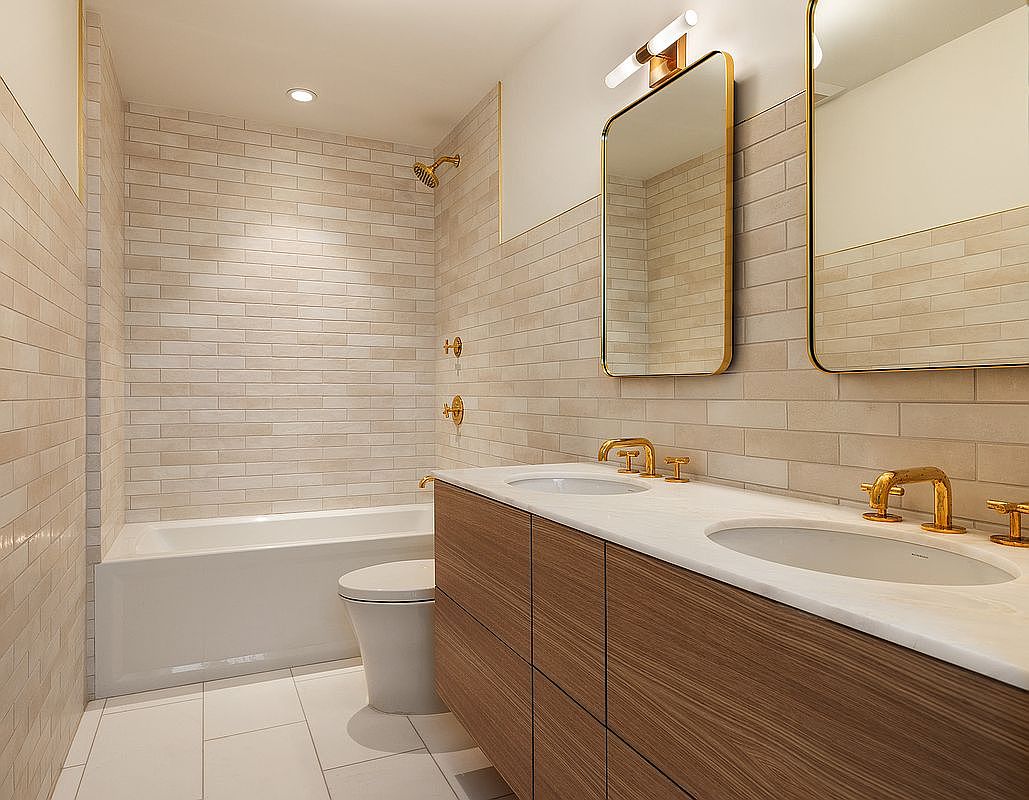 This modern primary bathroom features a sleek double vanity with wood-grain cabinetry, a white stone countertop, and gold-toned fixtures. The space is accented by floor-to-ceiling light-colored subway tiles, a deep soaking tub, and two minimalist mirrors with integrated lighting. The overall aesthetic is clean, contemporary, and luxurious, captured from a wide-angle perspective that highlights the efficient layout.