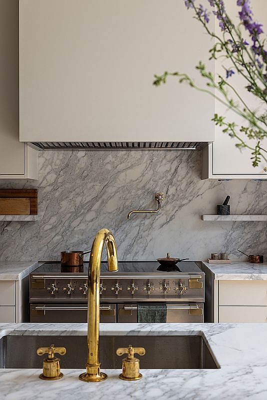 This elegant kitchen features a sophisticated blend of marble countertops and backsplash, complemented by a striking brass faucet and matching pot filler. A professional-grade stainless steel range sits beneath a minimalist range hood, creating a luxurious and functional culinary space. The perspective is a close-up, eye-level shot that emphasizes the high-end materials and refined design details.