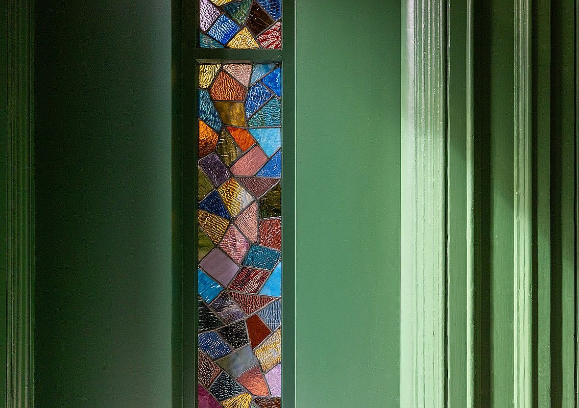 This close-up shot highlights a vibrant, multi-colored stained glass panel integrated into a wall painted in a rich, matte sage green. The glass features a mosaic of textured, irregular shards in hues of blue, amber, purple, and red, framed by classic, fluted wooden trim. The composition emphasizes the contrast between the organic, artistic glasswork and the structured, traditional architectural molding.