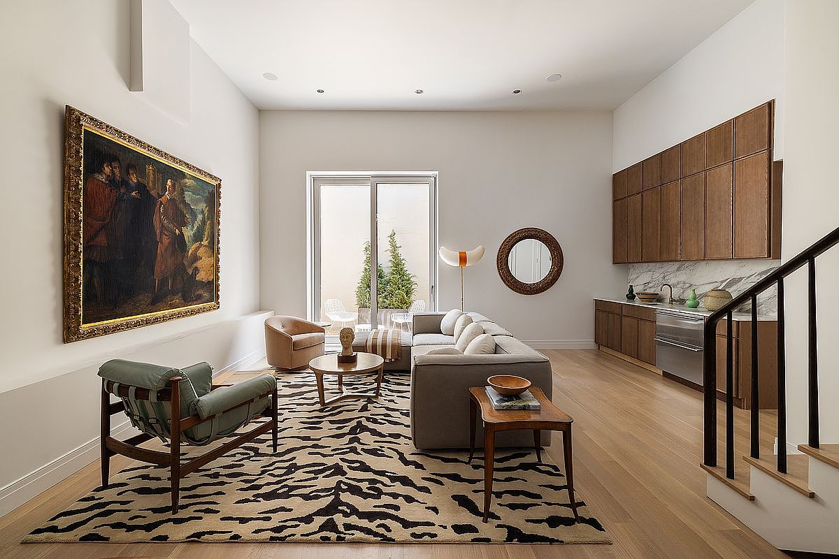 This sophisticated living room features a high-ceilinged, open-concept layout that seamlessly integrates a cozy seating area with a sleek, modern kitchenette. The space is anchored by a striking animal-print area rug, a large classical oil painting, and warm wood cabinetry, creating a refined blend of traditional and contemporary design. A large sliding glass door provides a view into a private outdoor patio, bathing the room in natural light and enhancing the airy, upscale atmosphere.