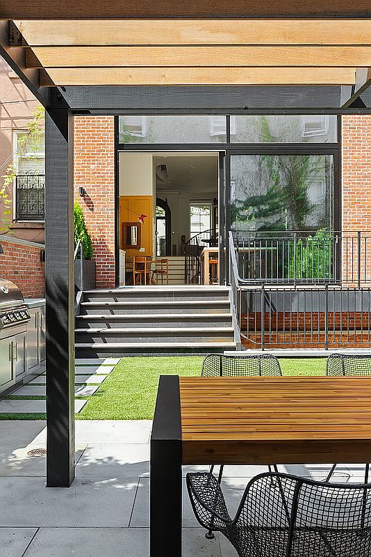 This image captures a modern, multi-level outdoor patio space featuring a wooden pergola, a sleek dining set, and a built-in stainless steel grill. The perspective looks from the patio toward a set of stairs leading up to a glass-walled entrance, revealing a glimpse into a stylish interior with warm yellow accents. The combination of brick architecture, manicured artificial turf, and contemporary metal furniture creates a sophisticated and inviting urban outdoor living area.