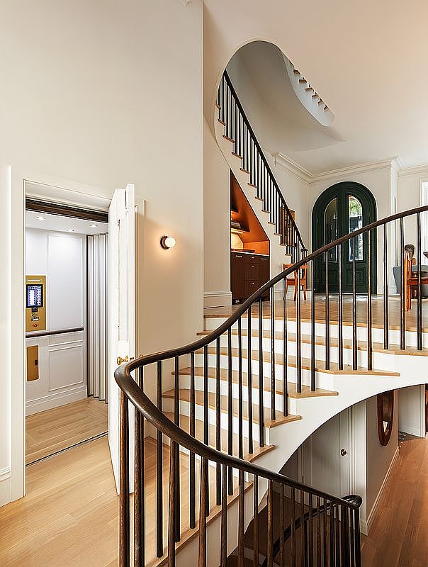 This elegant interior space features a grand, curved staircase with dark wooden railings and light wood treads, serving as the focal point of the entryway. To the left, a private residential elevator is integrated into the wall, while an arched doorway in the background leads to a bright, inviting living area. The neutral color palette and sophisticated architectural details create a luxurious and welcoming atmosphere.