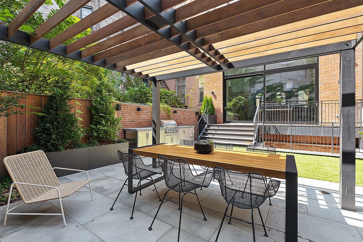 This sophisticated outdoor patio features a modern wooden pergola providing shade over a sleek dining set with wire-frame chairs. The space is complemented by a built-in stainless steel grill, lush greenery in contemporary planters, and a clean stone-tiled floor. The perspective captures a seamless transition from the outdoor dining area to the home's rear entrance, highlighting a refined and inviting urban oasis.
