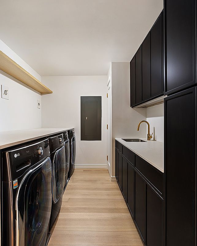 This modern, high-end laundry room features sleek black cabinetry paired with light wood flooring and a clean white countertop. The space is equipped with a row of LG appliances and a convenient sink, creating a functional yet stylish utility area. The perspective is a straight-on shot looking down the narrow corridor, emphasizing the efficient layout and contemporary design.