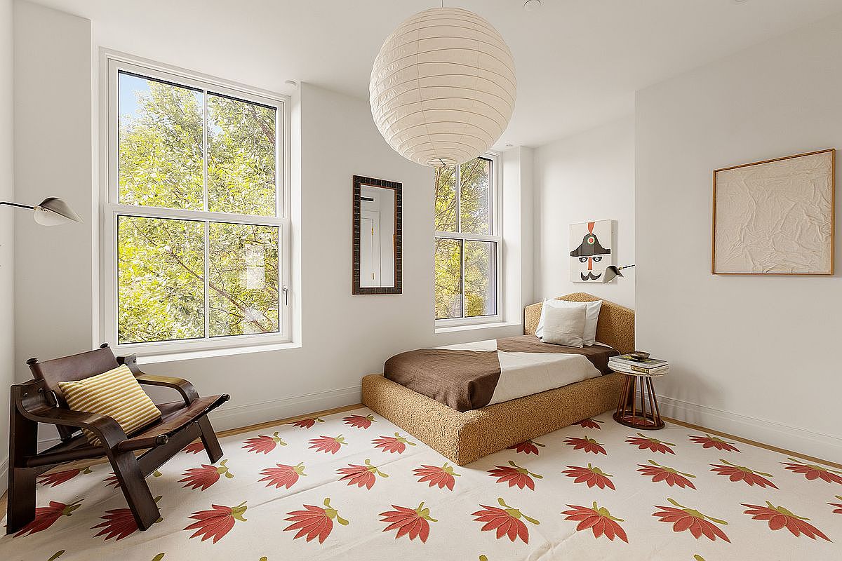 This bright and airy guest bedroom features a minimalist aesthetic with a textured, neutral-toned bed frame and a unique leaf-patterned area rug. The room is illuminated by a large paper lantern pendant light and natural light streaming through two tall windows, creating a serene and inviting atmosphere. A dark wood accent chair and a small side table add functional, stylish elements to the space.
