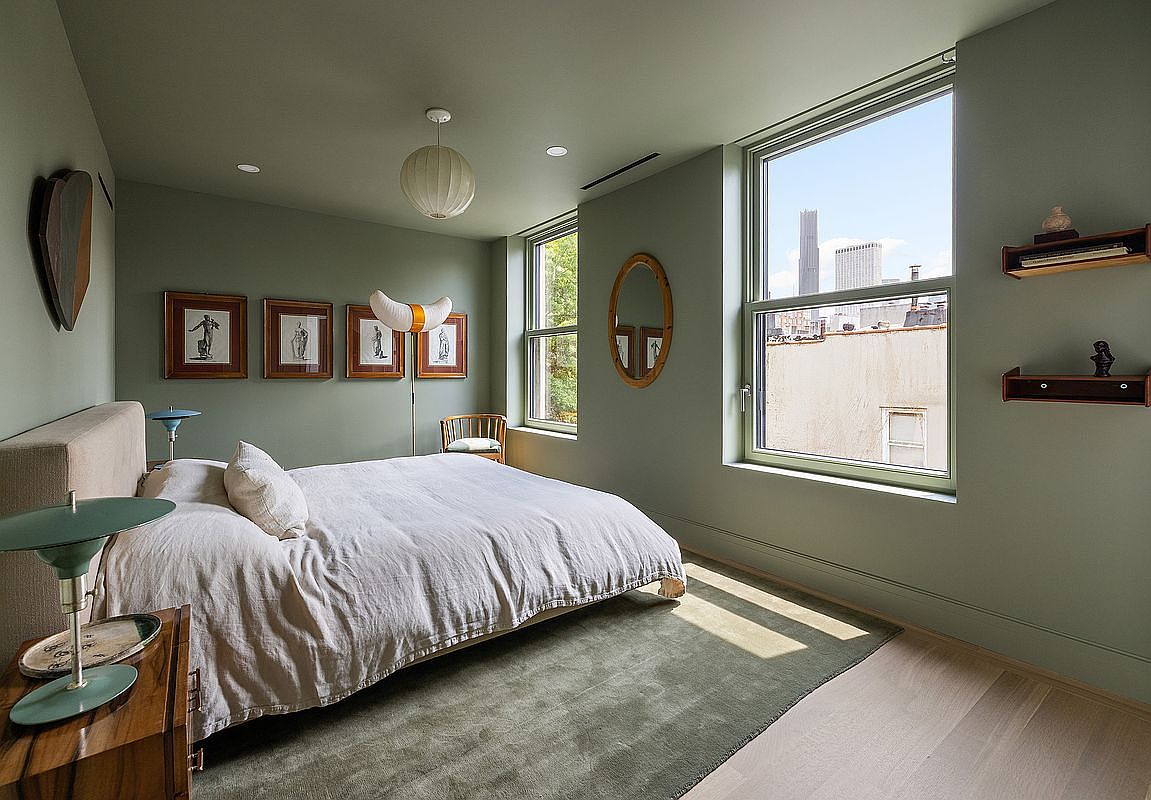 This serene primary bedroom features a calming sage green color palette, a plush bed with neutral linens, and a large area rug that anchors the space. The room is bathed in natural light from two large windows, one of which offers a clear view of the city skyline, while curated wall art and mid-century modern accents add a sophisticated touch. The perspective is a wide-angle shot from the corner of the room, capturing the tranquil atmosphere and elegant design.