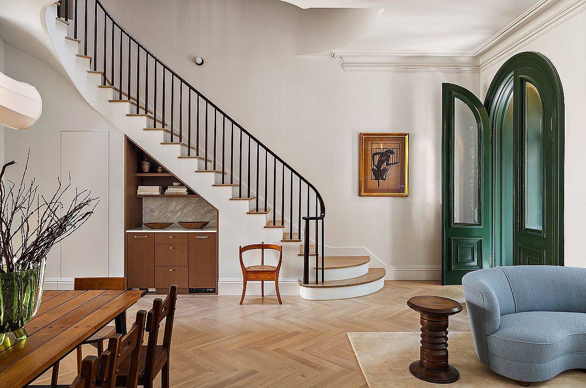 This elegant interior space features a grand staircase with a dark iron railing, complemented by a custom built-in wooden bar area tucked neatly underneath. The room showcases sophisticated design elements, including herringbone hardwood flooring, a curved light blue sofa, and a striking arched green doorway. The perspective is a wide-angle shot that captures the seamless blend of classic architectural details and modern, refined furnishings.