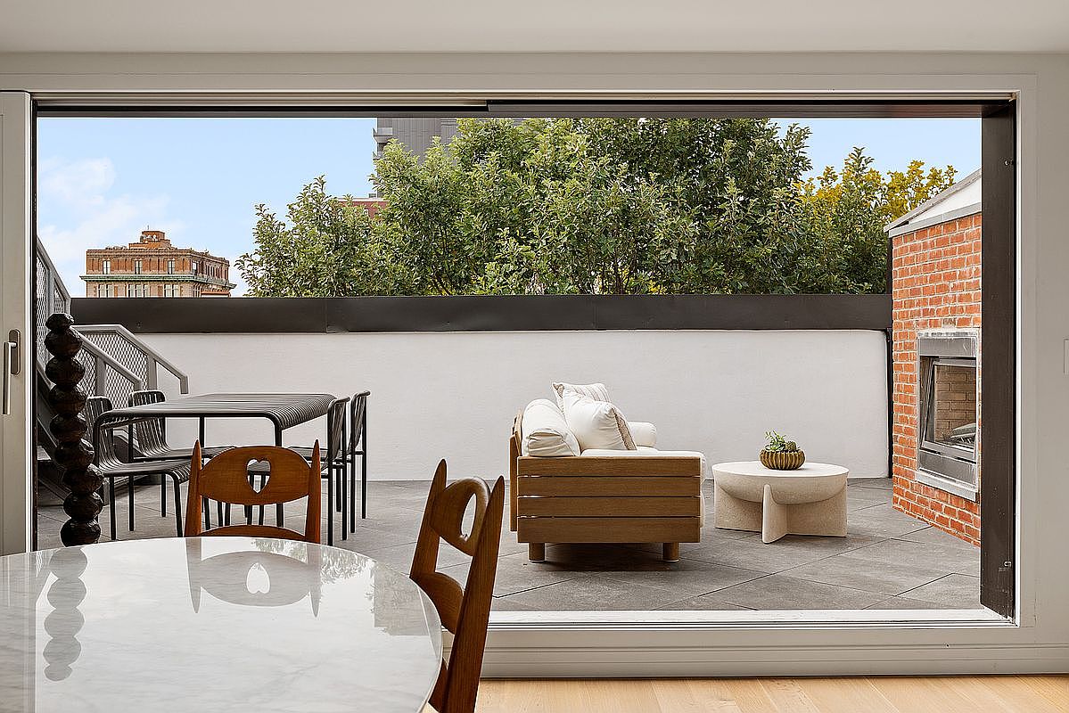 This image captures a seamless transition from an indoor dining area to a spacious, modern outdoor terrace. The balcony features a comfortable wooden sofa, a minimalist stone coffee table, and a dining set, all set against a backdrop of a brick fireplace and lush green trees. The perspective is framed from inside looking out, emphasizing the open-concept flow and the inviting, airy atmosphere of the space.