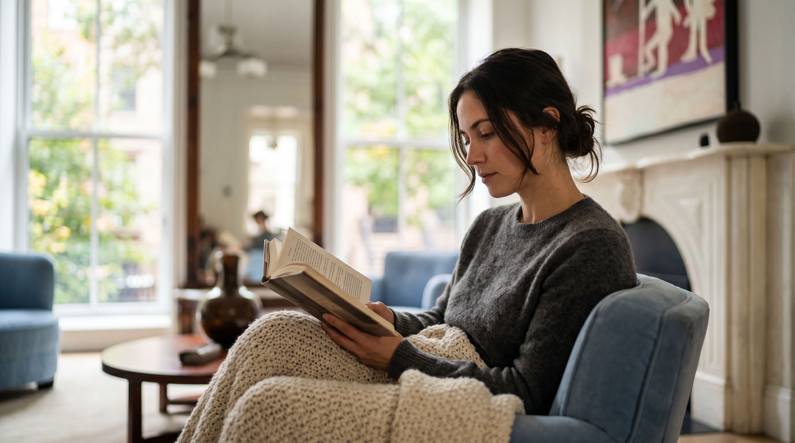 This living room area features a cozy and inviting atmosphere, centered around a comfortable blue armchair and a plush, knitted throw blanket. The room is accented by a classic fireplace mantle and large windows that provide abundant natural light, creating a bright and airy living space. The neutral color palette and tasteful decor contribute to a sophisticated, homey sense of comfort.