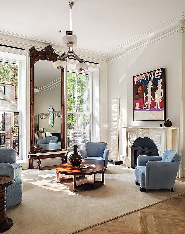 This elegant living room features a sophisticated blend of classic and contemporary design, anchored by a large, ornate wooden mirror and a marble fireplace. The space is furnished with plush, light blue armchairs arranged around a circular wooden coffee table, all set atop a neutral area rug on herringbone hardwood floors. Large windows flood the room with natural light, highlighting the high ceilings and the striking 'KANE' artwork above the mantel.
