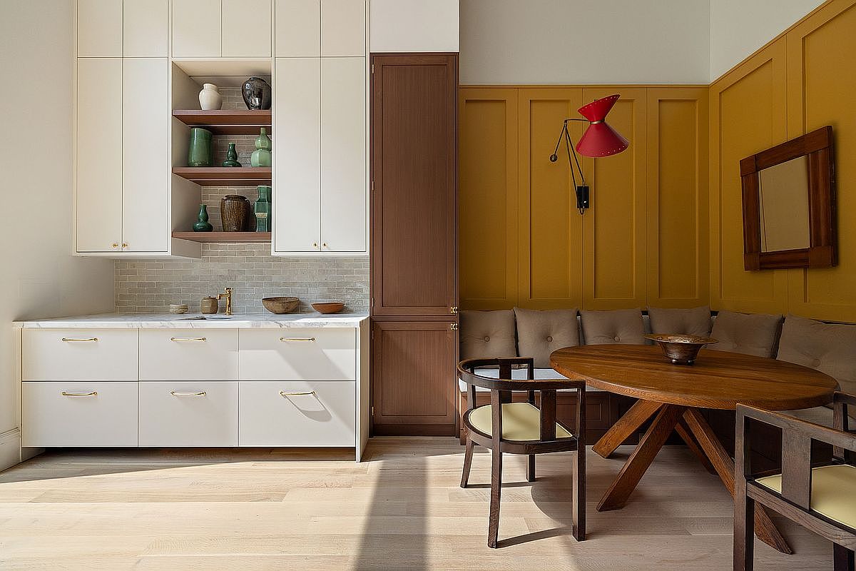 This sophisticated kitchen and dining nook features a seamless blend of modern white cabinetry and a warm, mustard-yellow paneled accent wall. A round wooden dining table is paired with a built-in banquette, creating an inviting breakfast area adjacent to a sleek wet bar with marble countertops. The space is illuminated by a striking red wall-mounted lamp, adding a vibrant pop of color to the refined, contemporary aesthetic.