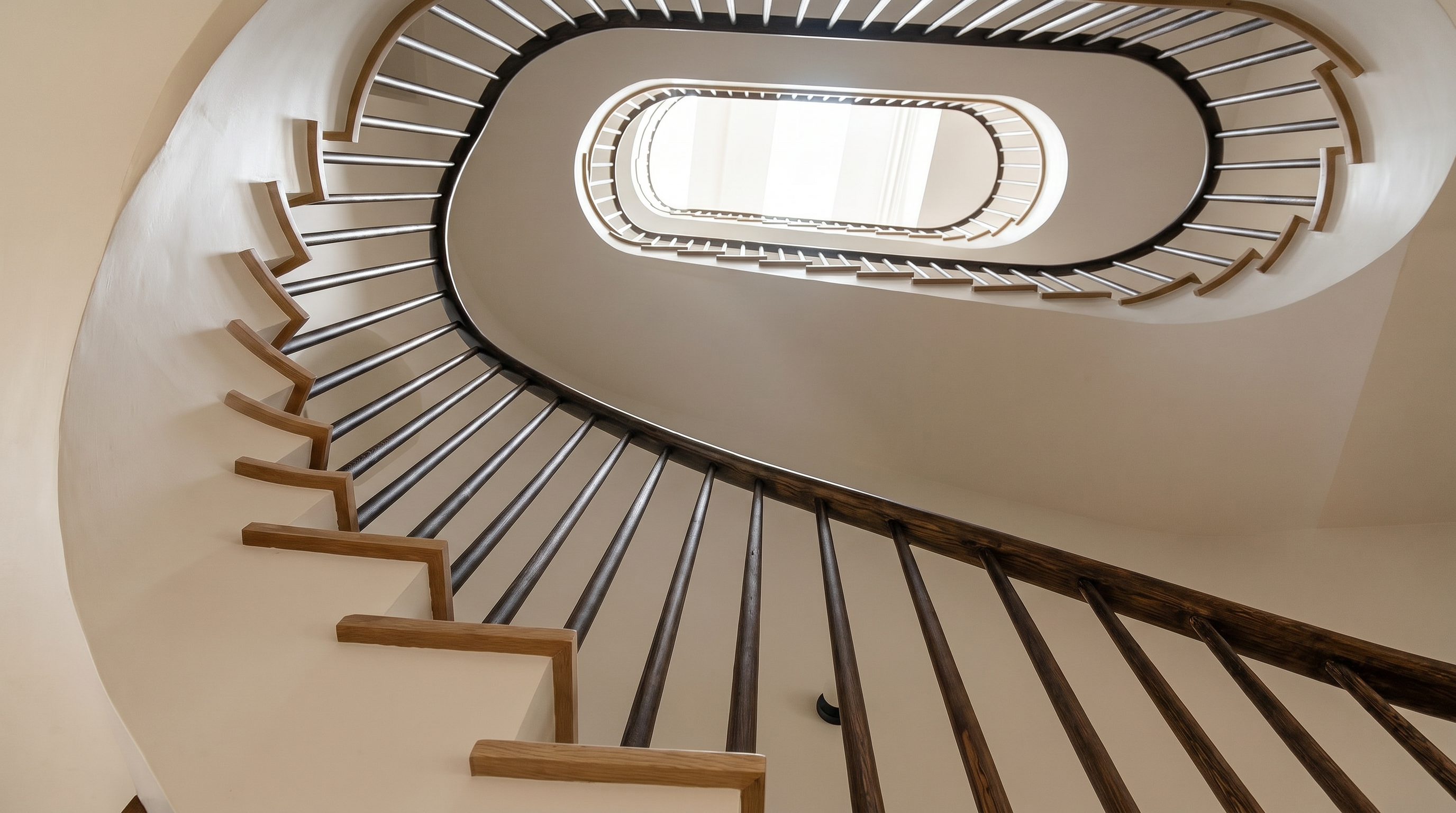 This image captures a striking, low-angle perspective of a modern, curved staircase that spirals upward toward a bright, oval-shaped skylight. The design features minimalist wooden treads, dark vertical balusters, and smooth, light-colored walls that emphasize the architectural geometry of the space. The clean lines and natural light create an elegant, airy, and sophisticated atmosphere.