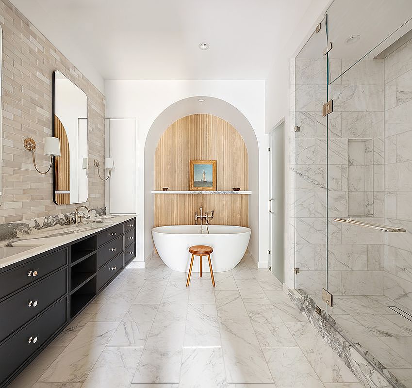 This luxurious primary bathroom features a striking arched alcove housing a freestanding soaking tub, accented by warm wood-slat paneling and a marble shelf. To the left, a long, dark-toned vanity with dual sinks and a large mirror provides ample storage, while a spacious glass-enclosed marble shower sits to the right. The room is finished with elegant marble flooring, creating a sophisticated and serene spa-like atmosphere.