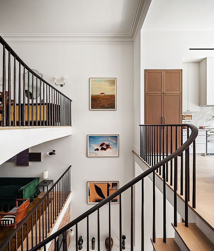 This sophisticated multi-level interior features a striking staircase with dark, slender railings that create a rhythmic, architectural focal point. The walls are adorned with a curated gallery of framed art, while the open-concept design allows for a seamless view between the upper landing and the lower living area. The space exudes a modern, minimalist aesthetic with clean lines, warm wood flooring, and high-end finishes.