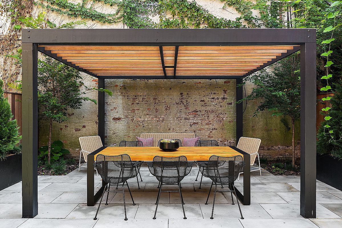 This elegant outdoor dining area features a modern pergola with a wooden slat roof, providing a sophisticated shaded space for entertaining. A sleek, contemporary dining table with black wire-frame chairs sits on a clean stone-tiled patio, set against a rustic, weathered brick wall covered in climbing vines. The scene captures a serene and stylish urban garden retreat, perfect for al fresco dining.