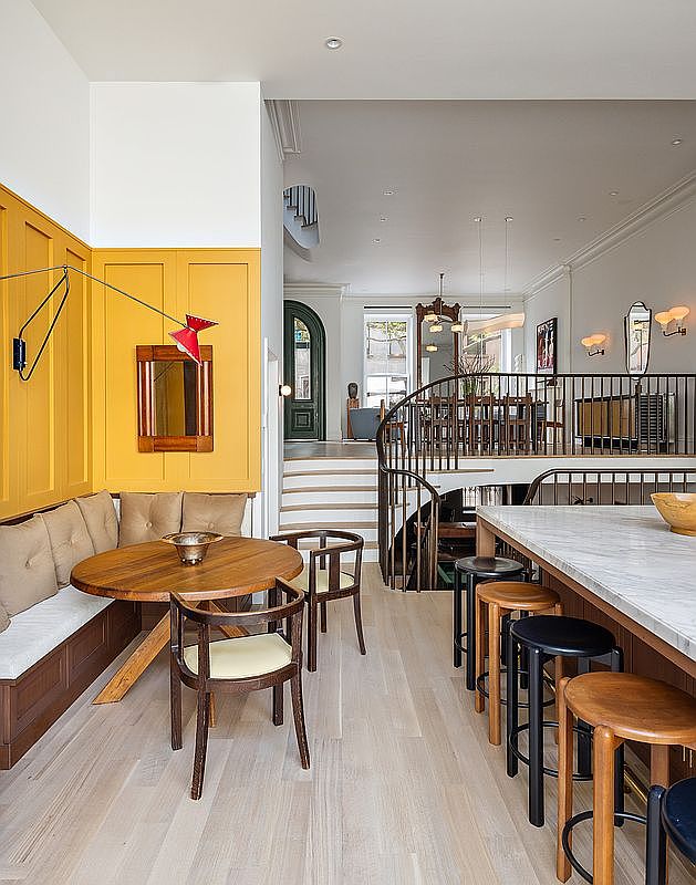 This bright, open-concept kitchen and dining area features a striking yellow paneled accent wall with a built-in banquette and a round wooden table. A marble-topped kitchen island with wooden stools provides a functional workspace, while a staircase leads to an elevated dining area in the background. The space blends modern design with warm, natural wood tones and elegant architectural details, creating an inviting and sophisticated atmosphere.