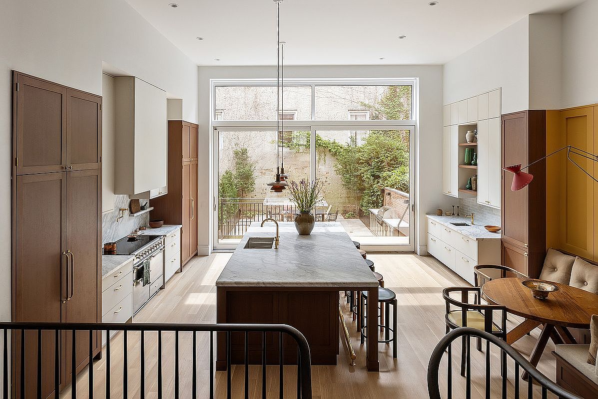 This sophisticated, high-end kitchen features a seamless blend of warm wood cabinetry and crisp white accents, centered around a massive marble-topped island. The space is bathed in natural light from floor-to-ceiling glass doors that open onto a lush, private garden patio. With its elegant pendant lighting, integrated appliances, and a cozy breakfast nook, the room offers a perfect balance of modern luxury and inviting warmth.