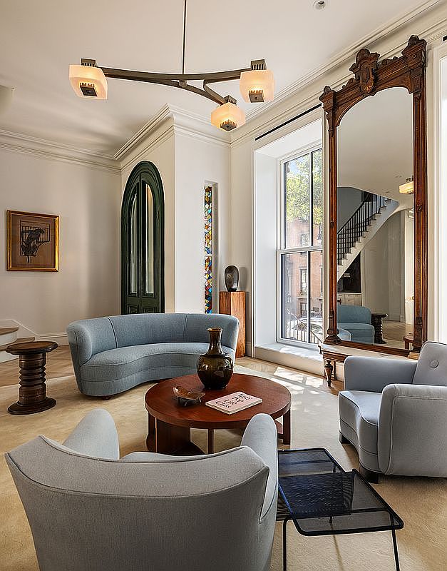 This elegant living room features a sophisticated blend of classic and contemporary design, highlighted by a curved light blue sofa and matching armchairs arranged around a circular wooden coffee table. A grand, ornate floor-to-ceiling mirror reflects the adjacent staircase, while a unique three-armed chandelier serves as a striking focal point above the seating area. The space is further enhanced by a tall, narrow stained-glass window and a dark arched doorway, creating a refined and artistic atmosphere.
