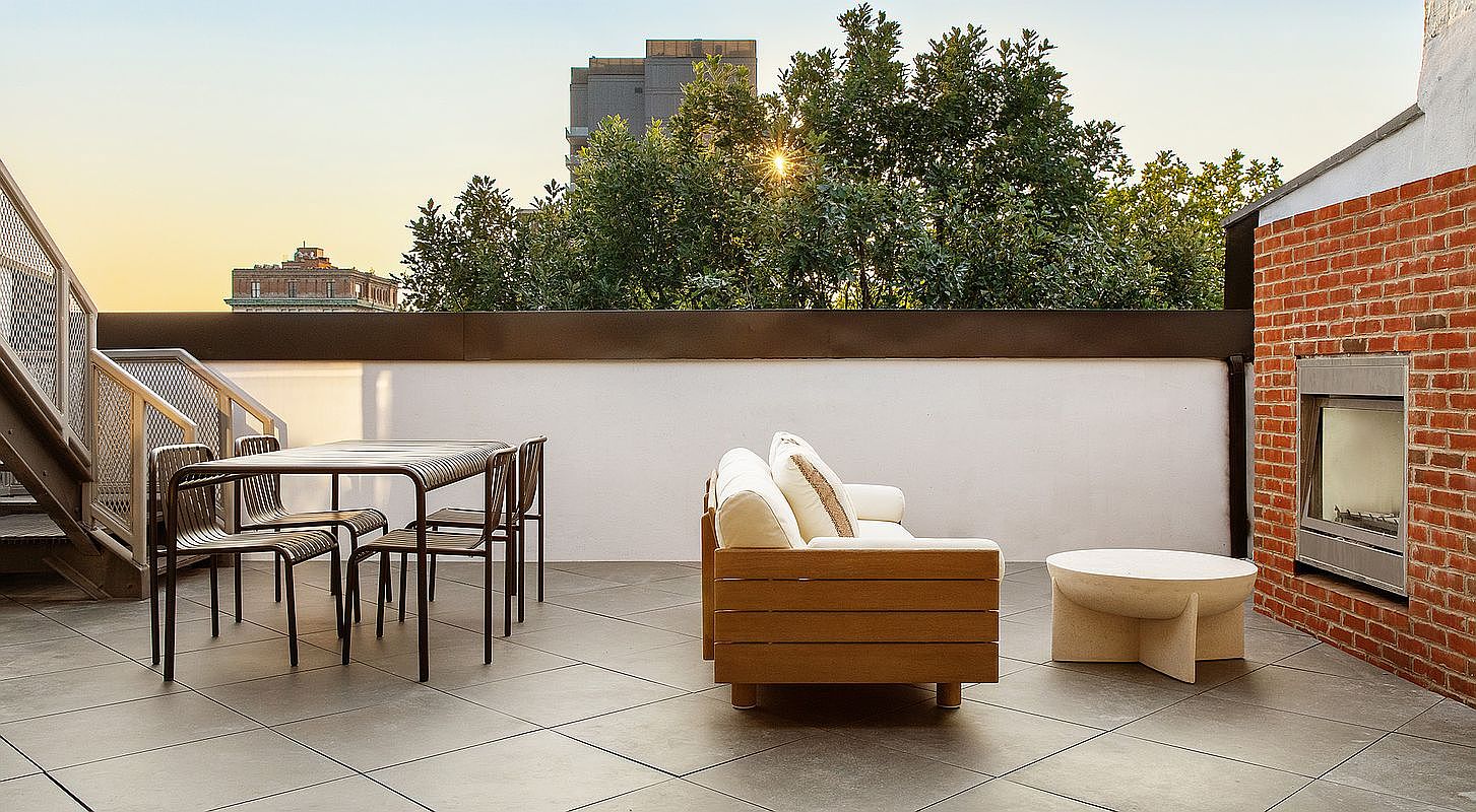 This spacious rooftop terrace features a modern outdoor dining set and a comfortable lounge area with a sofa and a unique stone coffee table. A built-in brick fireplace adds a cozy touch, while the open-air setting offers a scenic view of surrounding trees and urban architecture. The space is designed for relaxation and entertaining, blending contemporary furniture with classic brick elements.
