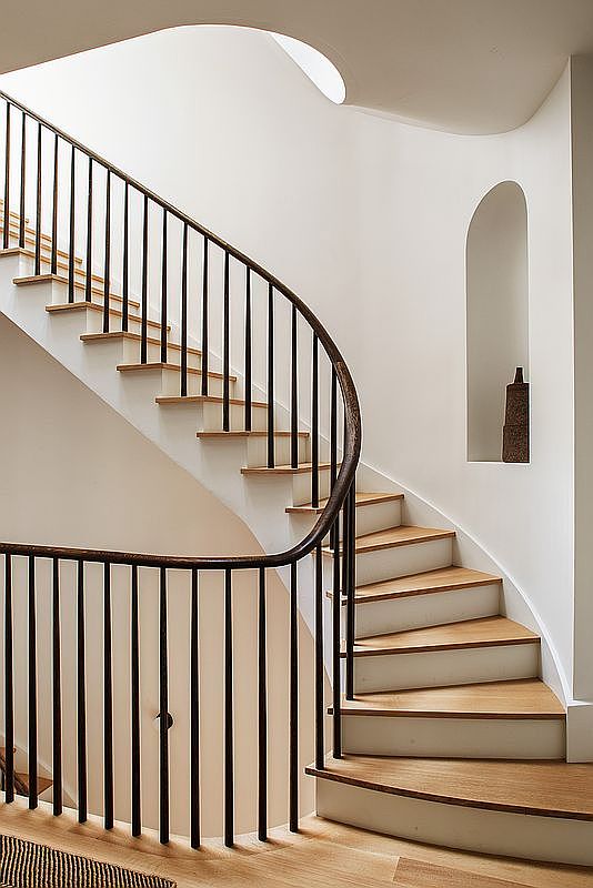 This elegant, minimalist staircase features a graceful curved design with light oak treads and crisp white risers. A dark, slender metal railing follows the curve, creating a striking contrast against the smooth, off-white walls. An arched wall niche adds a sophisticated architectural detail, housing a decorative sculptural piece that enhances the serene and modern aesthetic of the space.