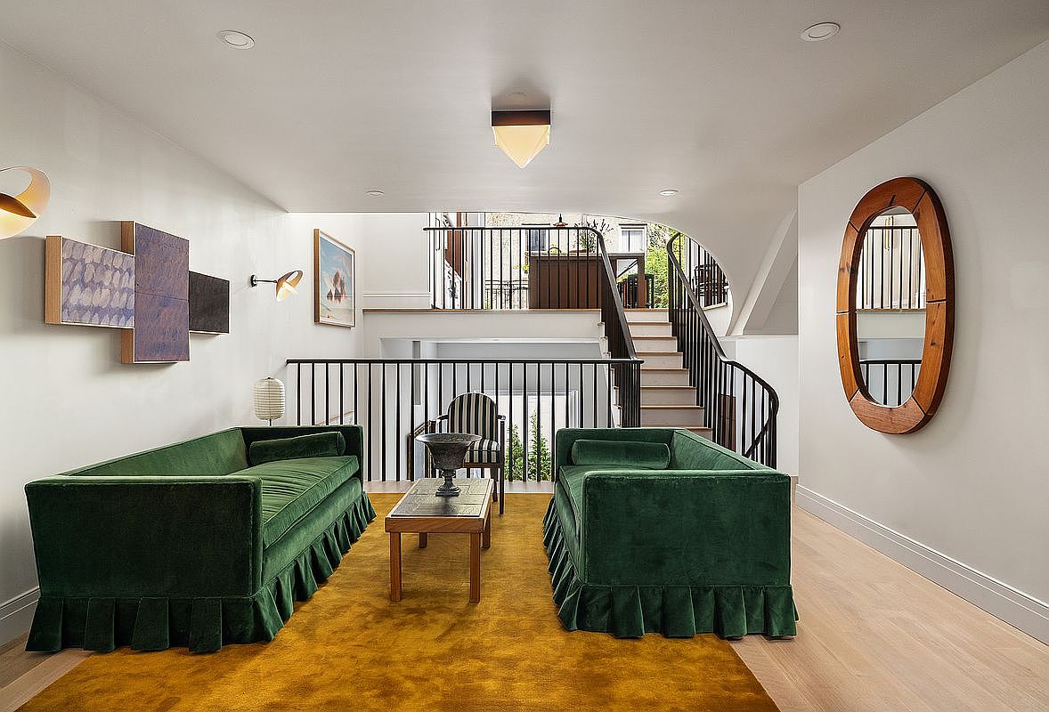 This elegant living room features two plush, emerald green velvet sofas with pleated skirts, arranged symmetrically around a wooden coffee table on a vibrant mustard yellow rug. The space is defined by a sophisticated, open-concept layout that overlooks a staircase with black iron railings, creating a sense of architectural depth and luxury. A large, oval-shaped wooden mirror on the right wall and abstract wall art on the left add a refined, artistic touch to the bright, neutral-toned interior.