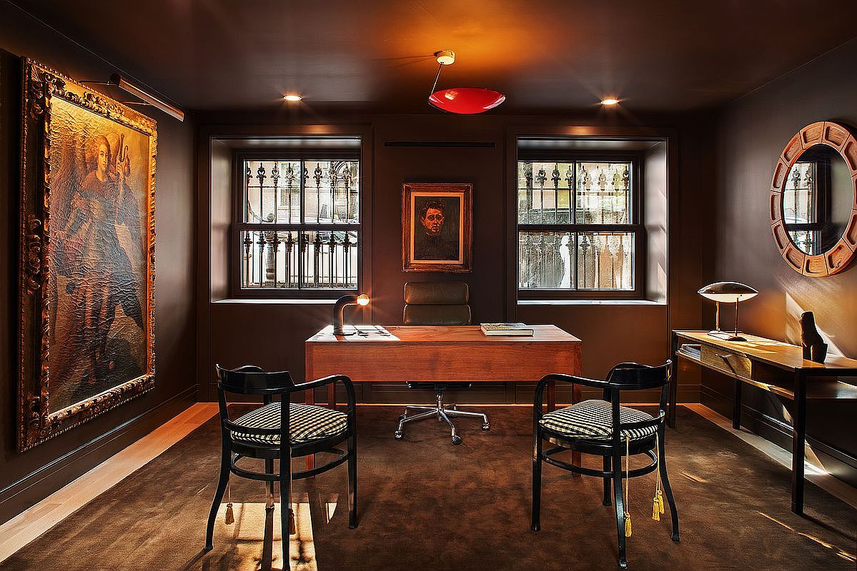 This sophisticated home office features a rich, dark color palette with deep brown walls and matching carpeting, creating an atmosphere of refined elegance. The room is anchored by a classic wooden desk, two patterned chairs, and a large, ornate framed painting, all illuminated by warm, focused lighting. The symmetrical layout, framed by two windows overlooking the exterior, provides a cinematic and professional aesthetic suitable for a high-end study.