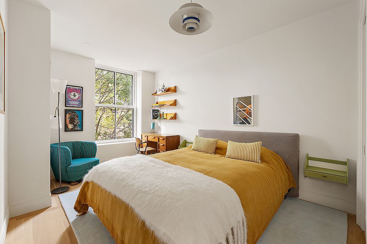 This bright and inviting bedroom features a large bed with a mustard-yellow duvet and a plush white throw, centered against a clean white wall. The room is accented by a vibrant teal striped armchair, a wooden desk area with floating shelves, and a large window that allows for ample natural light. The overall aesthetic is modern and cozy, with a balanced mix of textures and warm, cheerful colors.