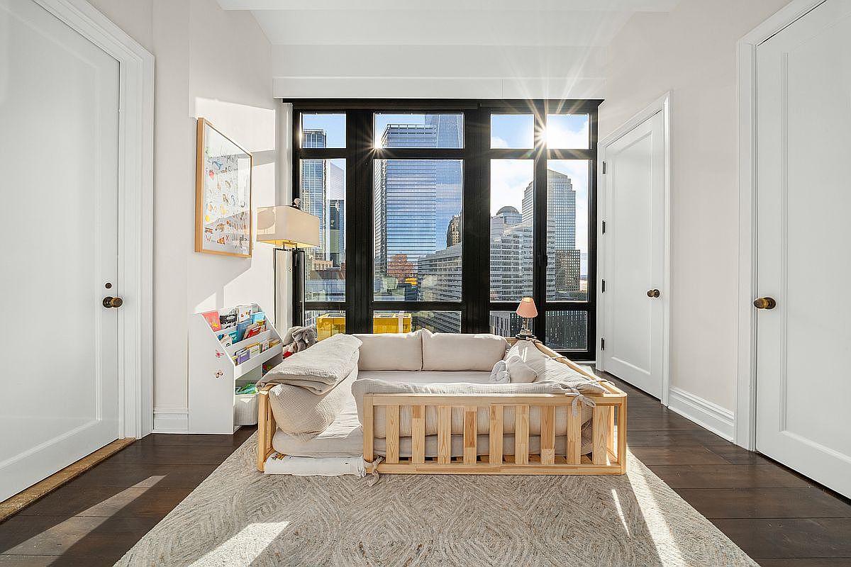 This bright and airy bedroom features a minimalist aesthetic with a low-profile wooden floor bed frame centered in the room. Large, floor-to-ceiling windows provide an expansive urban view of city skyscrapers, while a neutral-toned area rug and a small bookshelf add warmth to the space. The room is framed by two white doors, creating a clean, modern, and serene atmosphere.