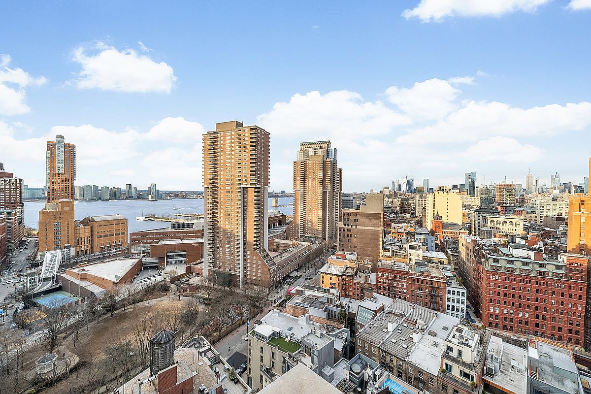This high-angle aerial view captures a sprawling urban cityscape, showcasing a dense mix of historic brick buildings and modern high-rise towers. The perspective highlights the proximity to a waterfront park and the Hudson River, emphasizing the vibrant, metropolitan lifestyle of the area. The bright, clear sky provides a panoramic backdrop that underscores the scale and architectural diversity of this prime city location.