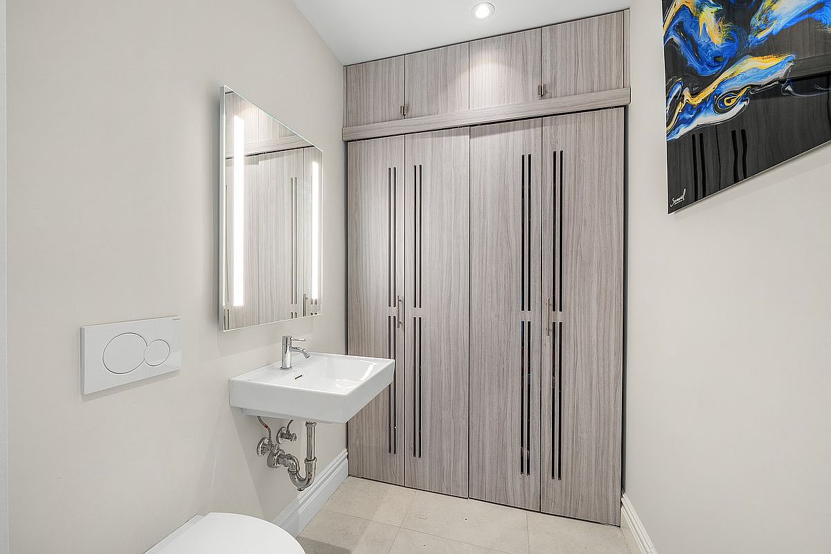 This modern powder room features a sleek wall-mounted sink with exposed chrome plumbing and a minimalist illuminated mirror. The space is anchored by floor-to-ceiling light wood-grain cabinetry that provides ample storage, complemented by a contemporary wall-mounted toilet flush plate. The overall aesthetic is clean, sophisticated, and highly functional, making it an ideal guest bathroom.