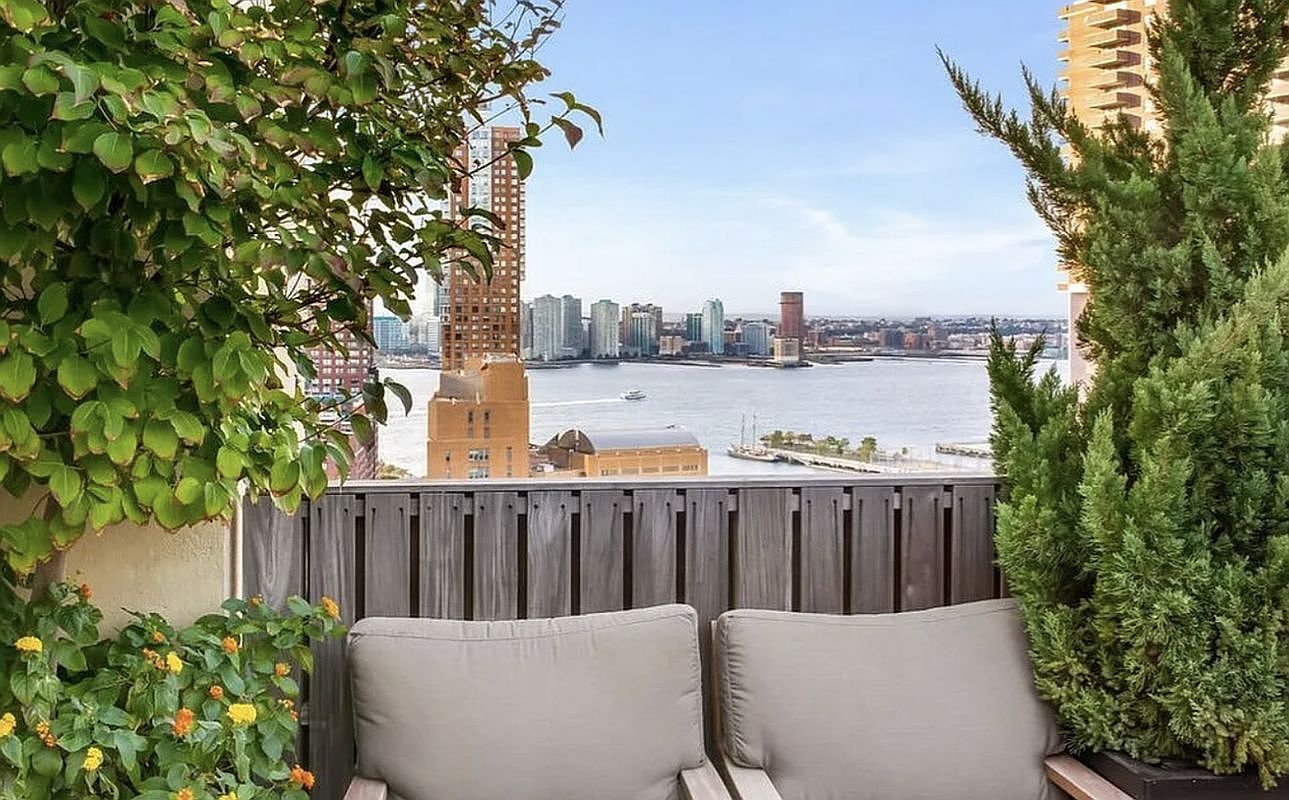 This image captures a serene balcony setting, featuring two comfortable chairs with plush cushions framed by lush greenery and a tall evergreen tree. The focal point is the stunning, unobstructed panoramic view of a river and a distant city skyline, creating an inviting and tranquil atmosphere. The perspective is from a seated position, emphasizing the private, relaxing nature of this outdoor space.
