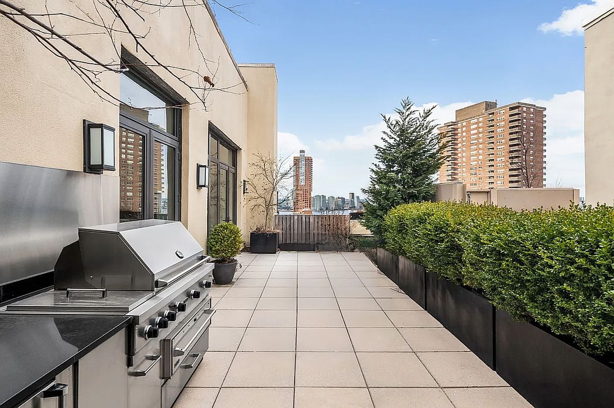 This expansive rooftop terrace features light-colored stone pavers and a built-in stainless steel grill, making it an ideal space for outdoor entertaining. A long, manicured hedge in a sleek black planter provides privacy and greenery, while the open layout offers stunning urban views of the city skyline. The perspective is a wide-angle shot from the terrace level, emphasizing the seamless transition between the indoor living space and the outdoor area.