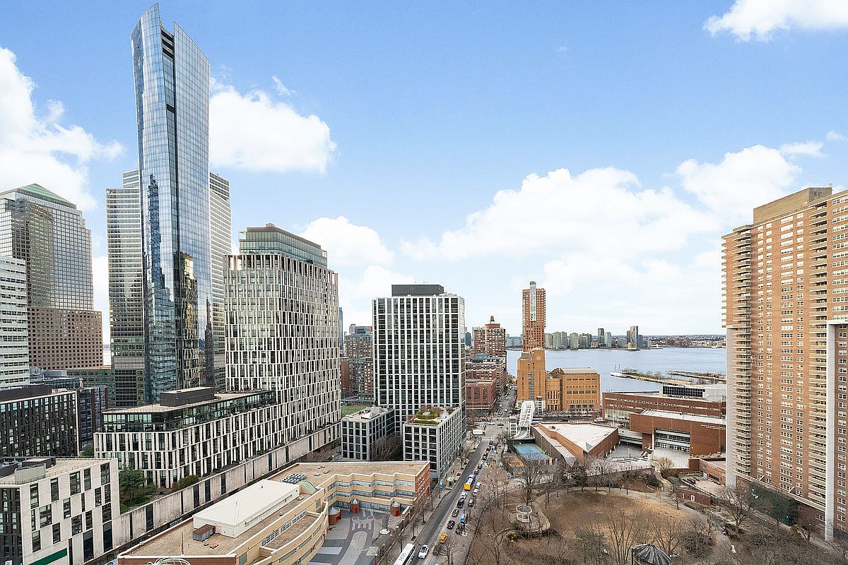 This high-angle aerial view captures a dense urban cityscape, dominated by a prominent, modern glass skyscraper that reflects the surrounding skyline. The perspective looks down onto a mix of contemporary high-rise residential buildings, a park area, and a bustling street leading toward a body of water in the distance. The scene conveys a sophisticated, metropolitan lifestyle with expansive views and a vibrant, active neighborhood setting.