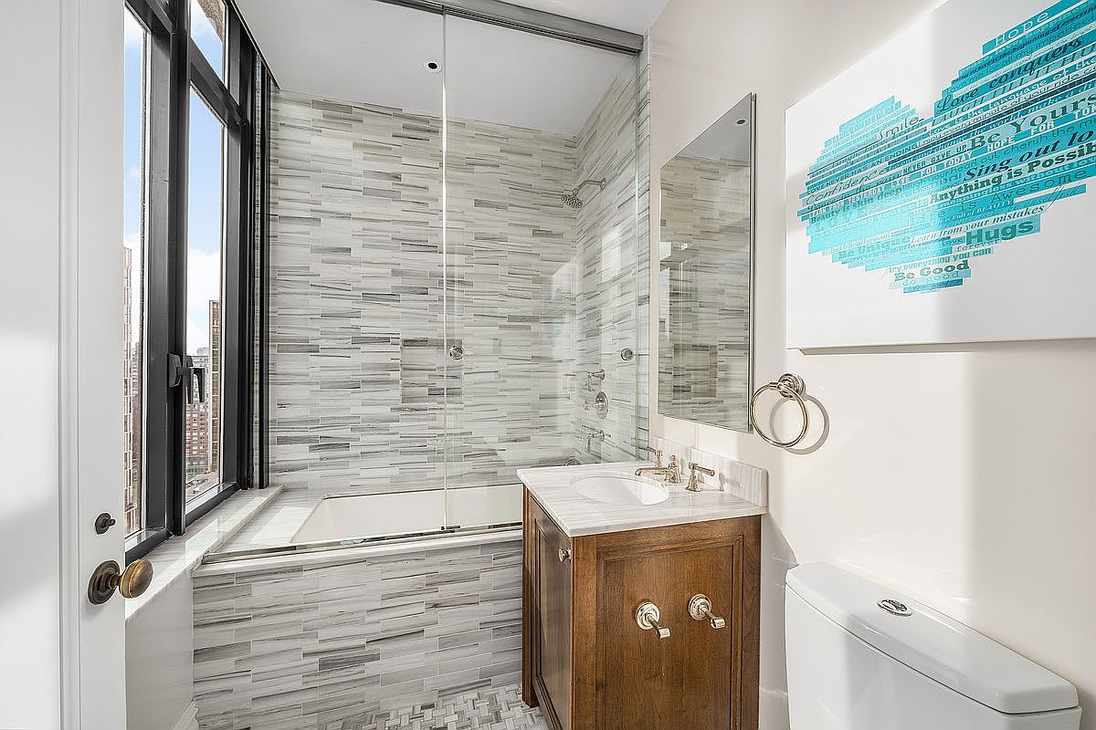 This compact, modern bathroom features a sleek glass-enclosed shower-tub combination with textured, horizontal-patterned wall tiles. A wooden vanity with a white stone countertop and a framed mirror adds warmth to the space, while a motivational art piece hangs on the wall above the toilet. The room is bright and functional, with a large window providing natural light and a view of the city skyline.