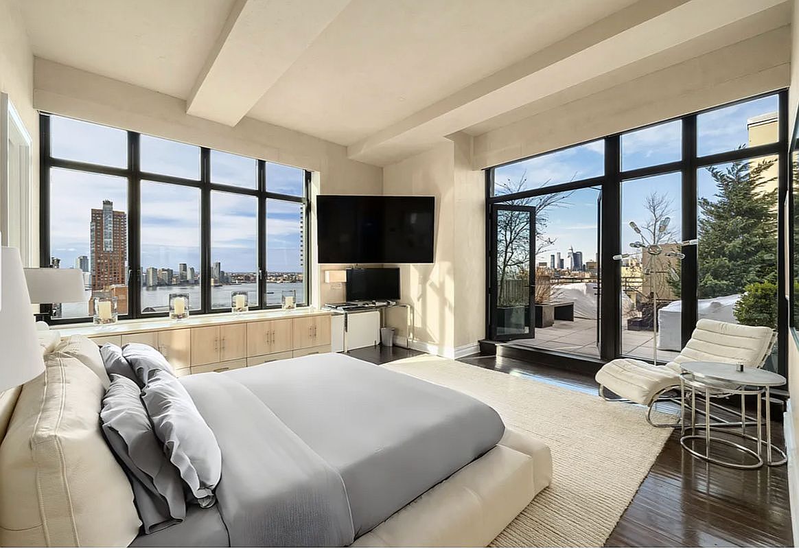 This luxurious primary bedroom features a sleek, modern aesthetic with a low-profile platform bed as the centerpiece. Expansive floor-to-ceiling windows wrap around the room, offering breathtaking panoramic city and waterfront views while flooding the space with natural light. A private terrace accessible through glass doors and a minimalist lounge area enhance the sophisticated, high-end urban living experience.