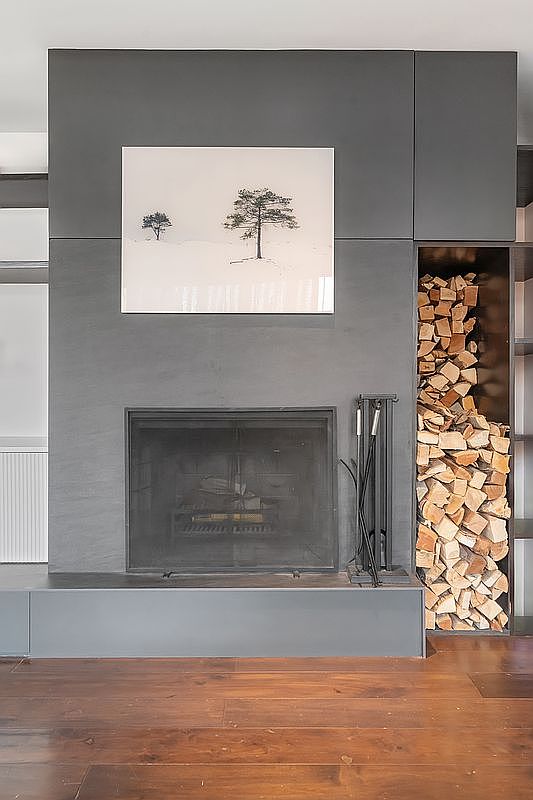 This interior shot features a striking, modern fireplace wall finished in a matte charcoal gray, serving as a bold focal point. A minimalist landscape print hangs above the fireplace, while a built-in vertical niche to the right provides organized, aesthetic storage for firewood. The warm, rich tones of the hardwood flooring create a sophisticated contrast against the dark, industrial-inspired fireplace structure.