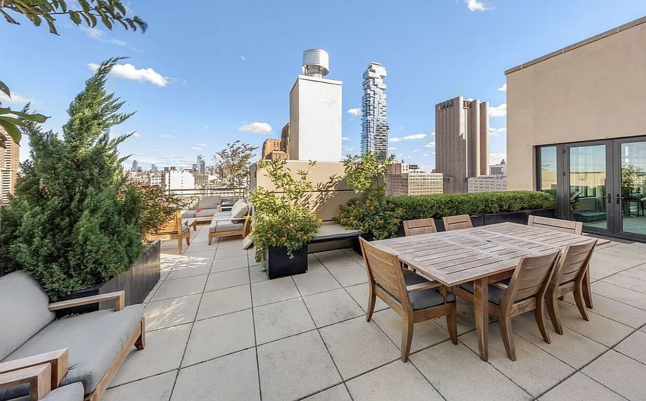 This expansive rooftop terrace offers a sophisticated outdoor living space with a wooden dining set and comfortable lounge seating. The area is framed by lush potted greenery and planters, providing a serene contrast to the dramatic urban skyline backdrop featuring iconic New York City architecture. The wide-angle perspective captures the seamless blend of private luxury and vibrant city life, making it an ideal setting for entertaining.