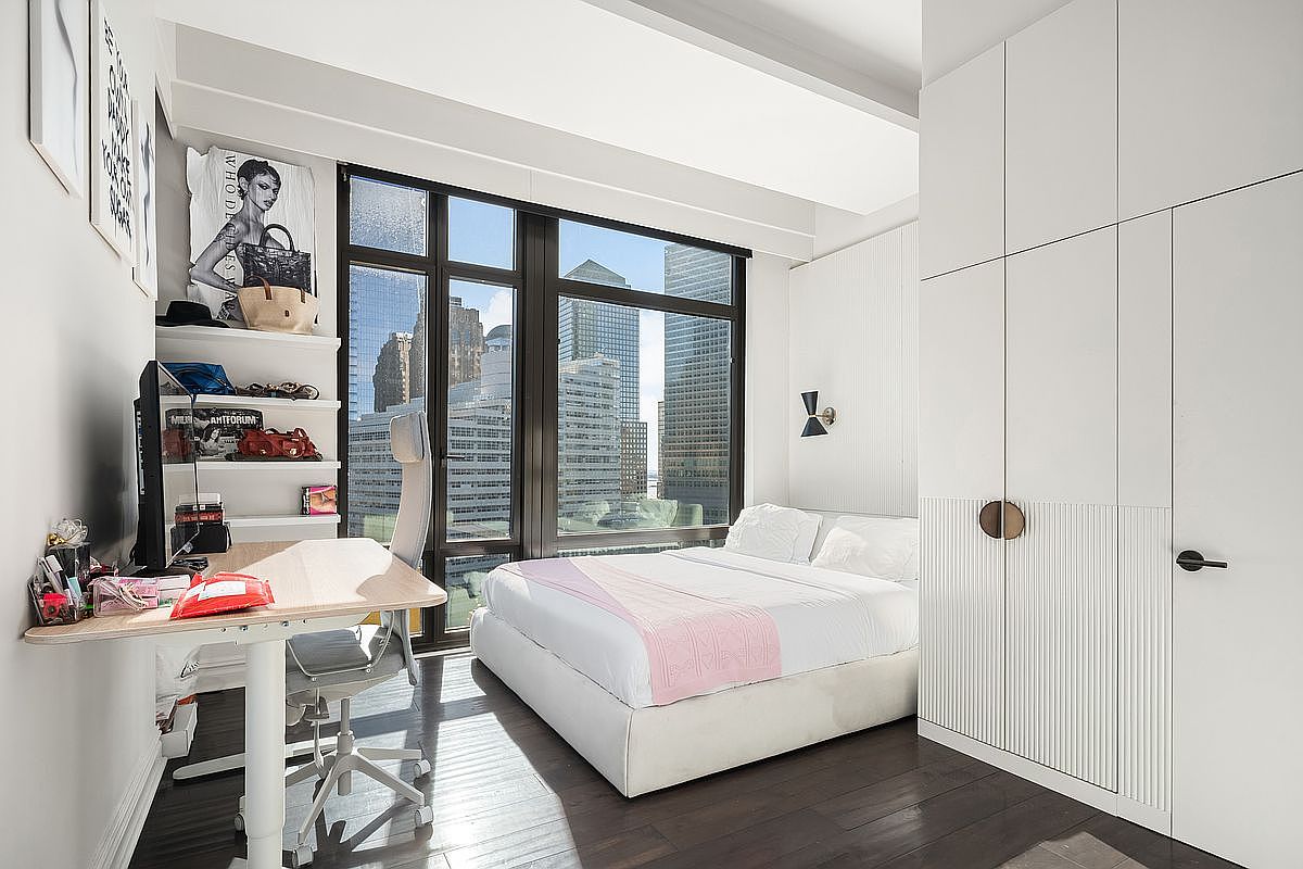 This modern, light-filled bedroom features a sleek, minimalist aesthetic with a queen-sized bed, a built-in desk workspace, and floor-to-ceiling white cabinetry. Large windows provide an expansive, impressive view of the city skyline, creating a bright and airy atmosphere. The dark wood flooring contrasts beautifully with the crisp white walls and furniture, offering a sophisticated urban living experience.
