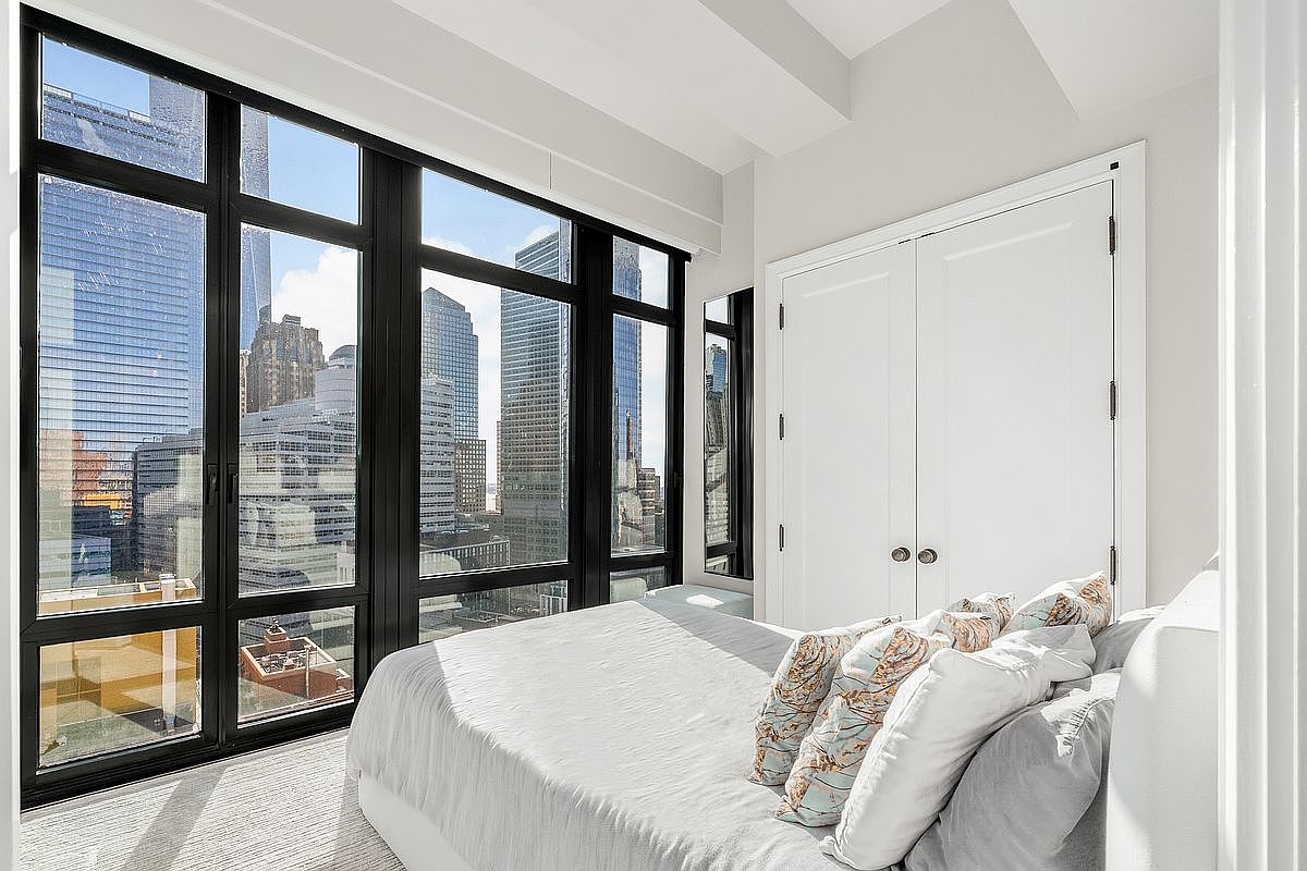 This modern bedroom features floor-to-ceiling windows that offer a stunning, expansive view of the city skyline. The room is furnished with a plush bed adorned with patterned pillows and a neutral-toned duvet, creating a serene and sophisticated atmosphere. A set of white double doors suggests a spacious closet, while the clean lines and bright, airy aesthetic emphasize the luxury urban living experience.