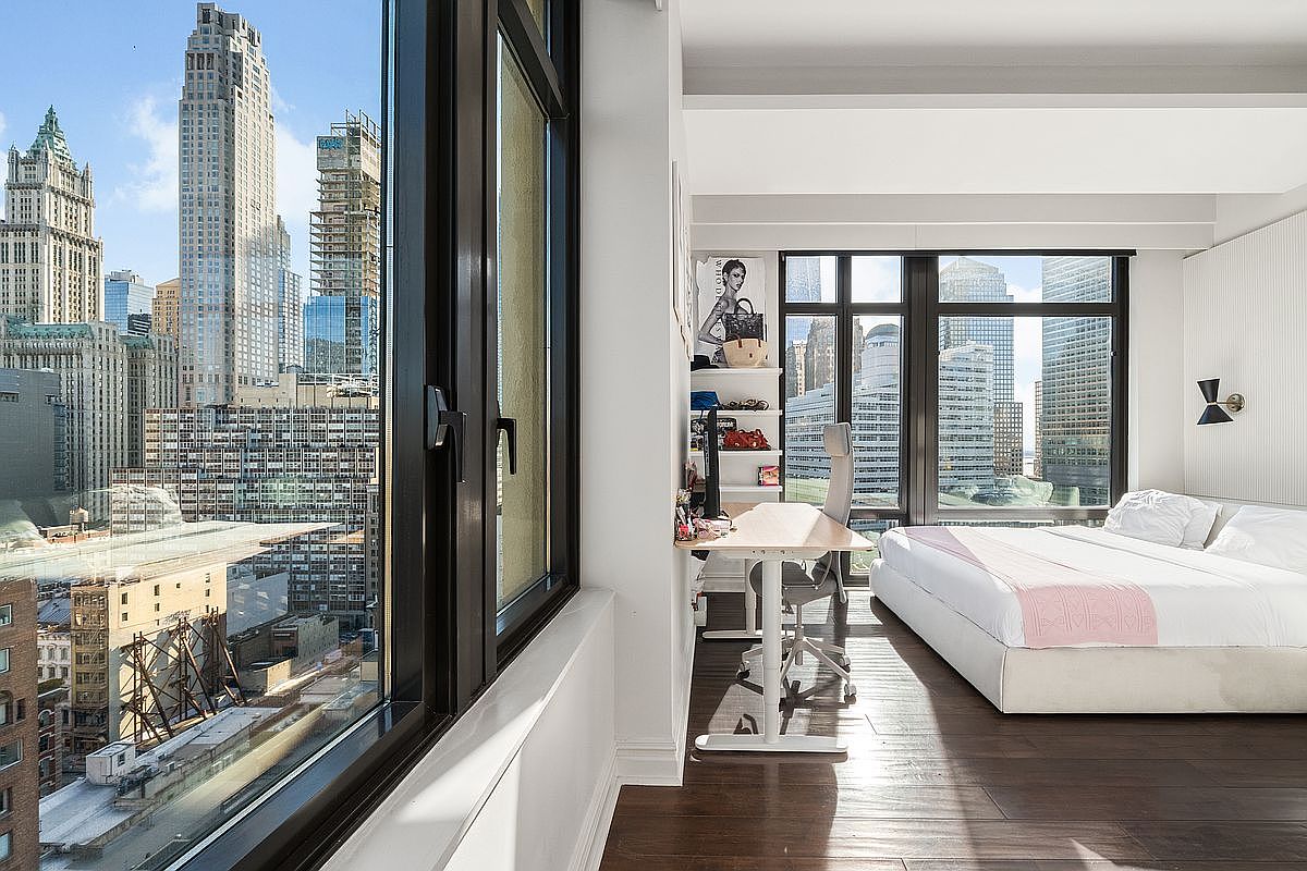 This modern, sun-drenched bedroom features a sleek white bed with a soft pink throw, positioned next to floor-to-ceiling windows that offer a dramatic, panoramic view of the New York City skyline. A minimalist standing desk setup is integrated into the space, creating a functional work-from-home area that overlooks the urban landscape. The room is characterized by its clean lines, dark hardwood flooring, and a sophisticated, high-rise aesthetic.