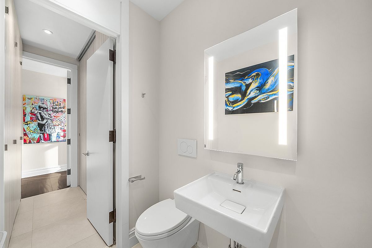 This modern powder room features a sleek, wall-mounted white sink and a minimalist toilet, complemented by a contemporary LED-backlit mirror. The space is accented by vibrant, artistic wall decor, including a colorful piece above the sink and a striking 'I will love you forever' print visible through the doorway. The neutral color palette and clean lines create a sophisticated and artistic atmosphere.
